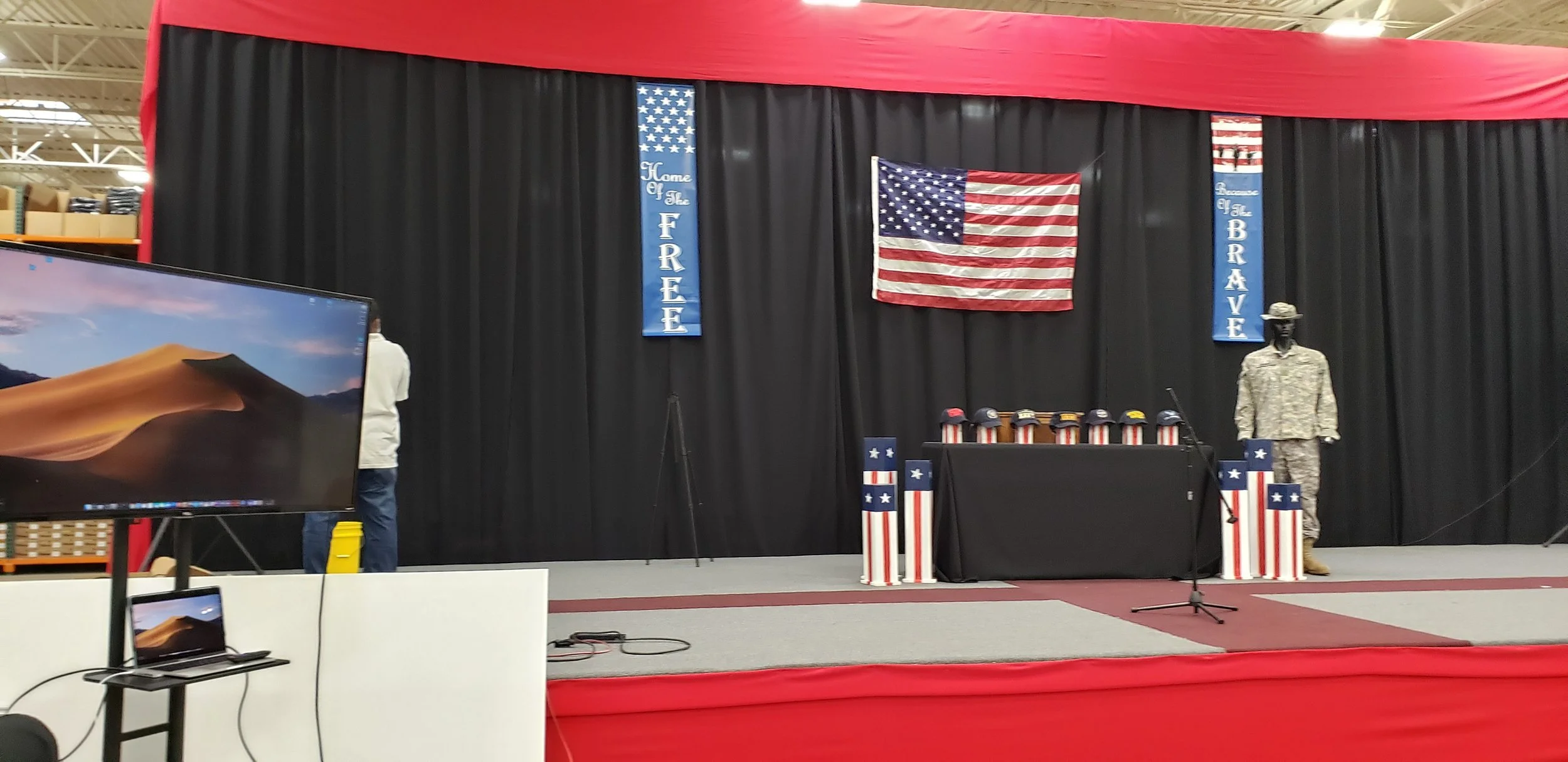 Stage decorated with patriotic American themes, featuring an American flag, table with hats, and mannequin dressed in camouflage military uniform, set against black curtains with blue banners reading 'Home of the Free' and 'Because of the Brave' in an indoor venue.
