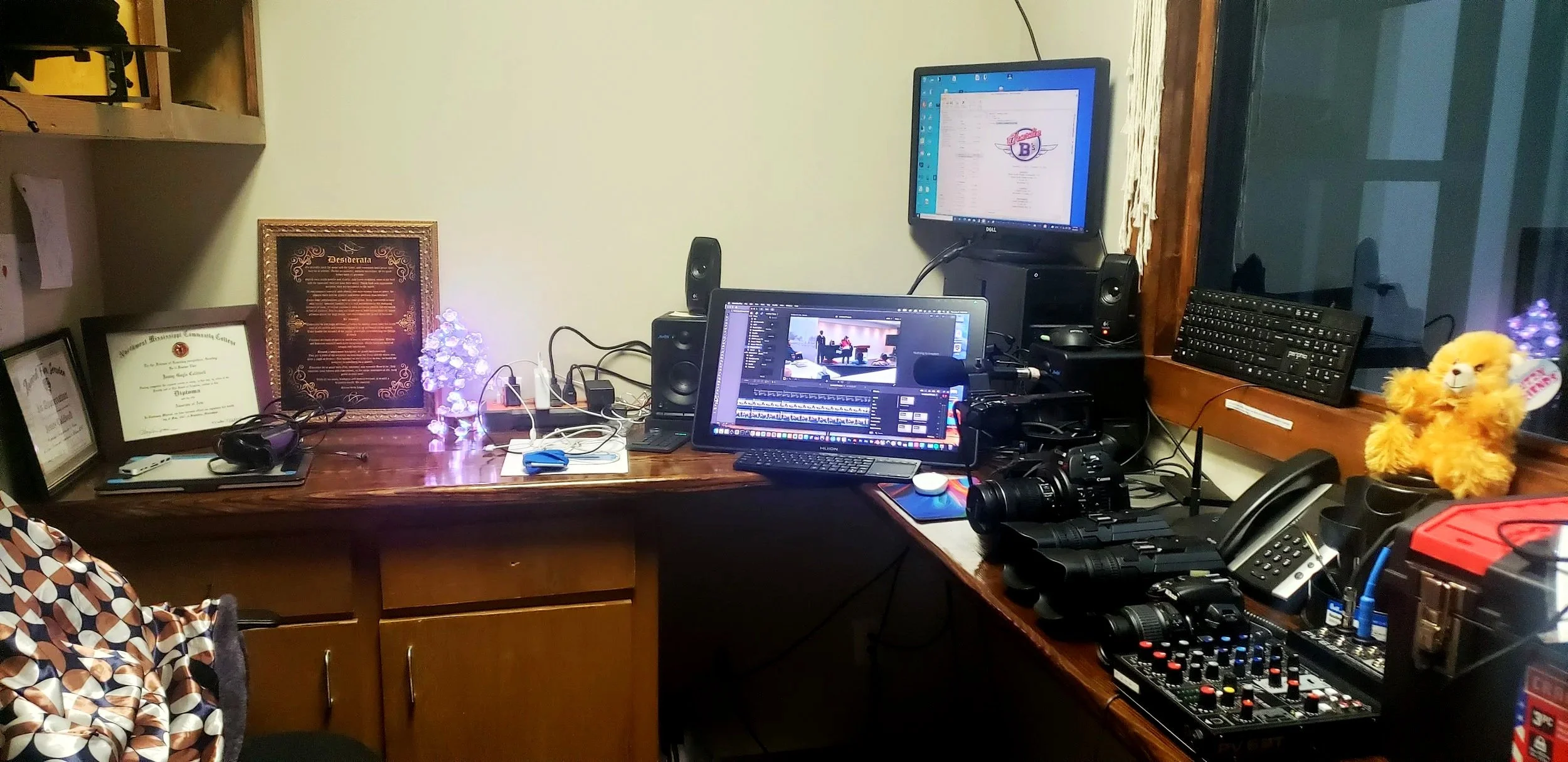 An office desk with multiple monitors, cameras, microphones, and various electronic equipment, with diplomas and certificates on the wall and a plush lion toy on the window sill.