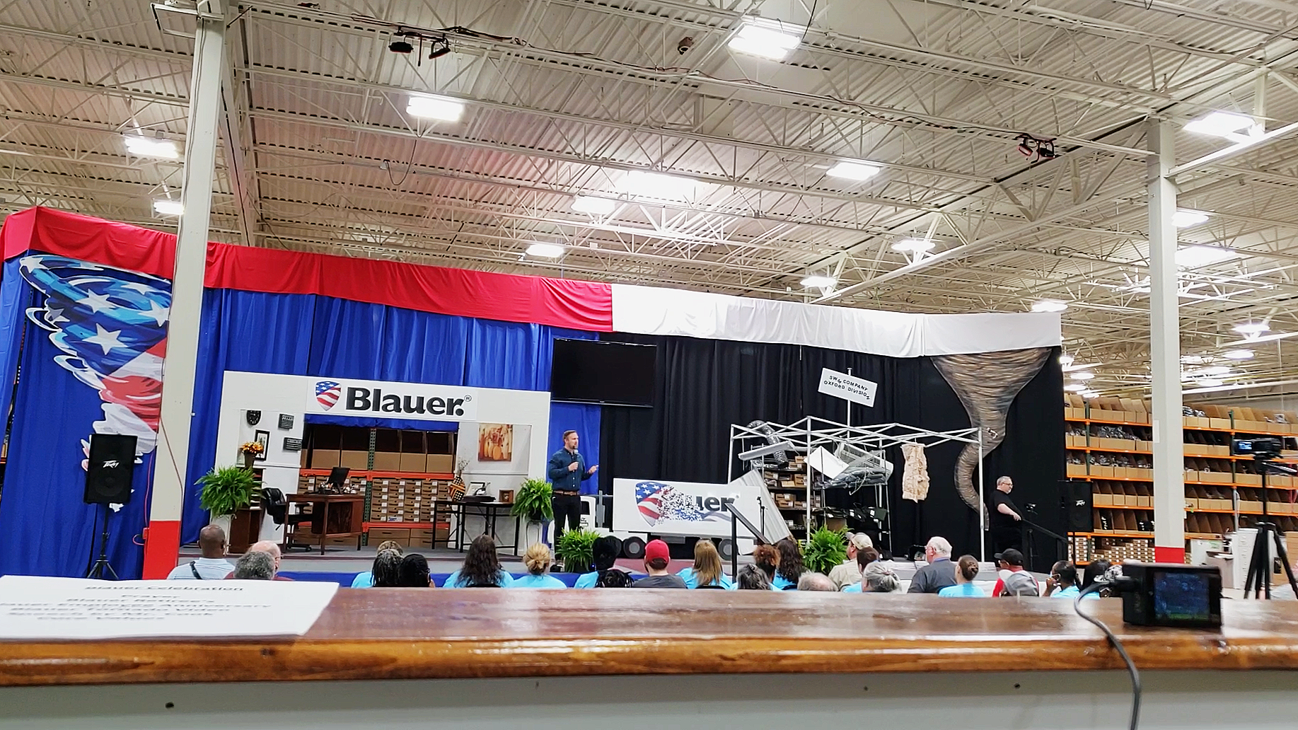 Presentation stage set up with a blue, red, white, and black backdrop. There are two speakers on each side of the stage, plants, a desk, and a microphone. Several people are seated in front, facing the stage. The event appears to be in a large warehouse or store.