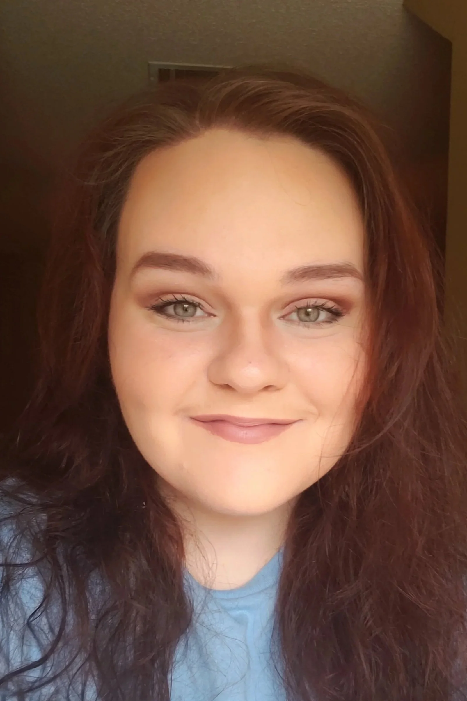 A close-up of a young woman with long, curly reddish-brown hair, light green eyes, and light makeup, smiling at the camera.