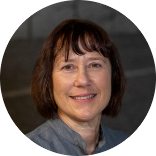 A circular profile image of a Vancouver Prosthetics & Orthotics employee, woman with short brown hair and a smile, wearing a gray top, posing in front of a gray background in Vancouver, BC.