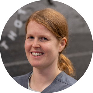 A circular profile image of a Vancouver Prosthetics & Orthotics employee, a woman with ginger hair in a ponytail smiling, wearing a gray jacket, standing in front of a gray background in Vancouver, BC.