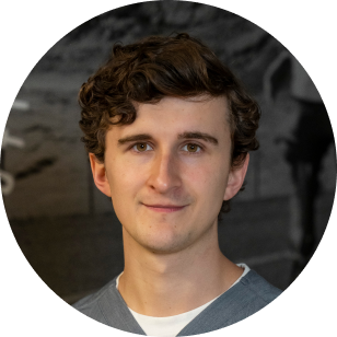 A circular profile image of a Vancouver Prosthetics & Orthotics employee, a young man with curly brown hair and fair skin, smiling softly, wearing a gray sweater, standing in front of a dark grey background in Vancouver, BC.