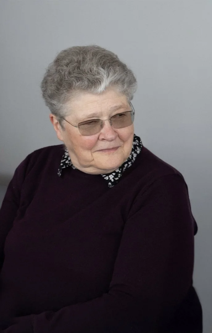 An elderly woman with gray curly hair and glasses, wearing a black sweater with a patterned collar, sitting against a plain gray background.