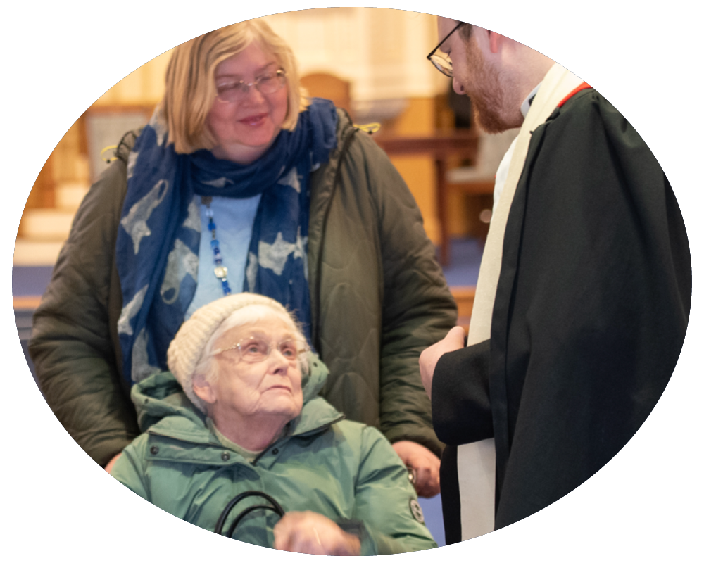 An elderly woman in a wheel chair being visited by a man in robes and an older woman in a green jacket, indoors.