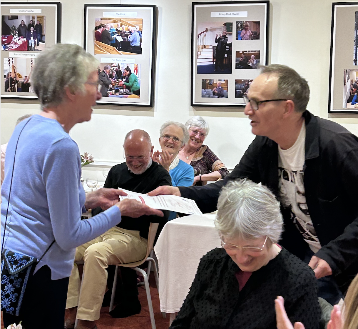 A woman in a light blue sweater is handing a paper or certificate to a man with glasses, smiling, at a gathering or celebration. Several seated and standing people are in the background, some smiling and clapping. The setting appears to be a restaurant or event hall with framed photographs on the wall.