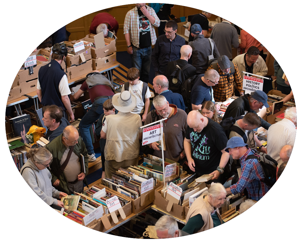 Crowd browsing books at an indoor book sale or market with tables and cardboard boxes of books, and signs indicating sections like 'Art', 'Architecture', 'History, Politics, Archaeology'.