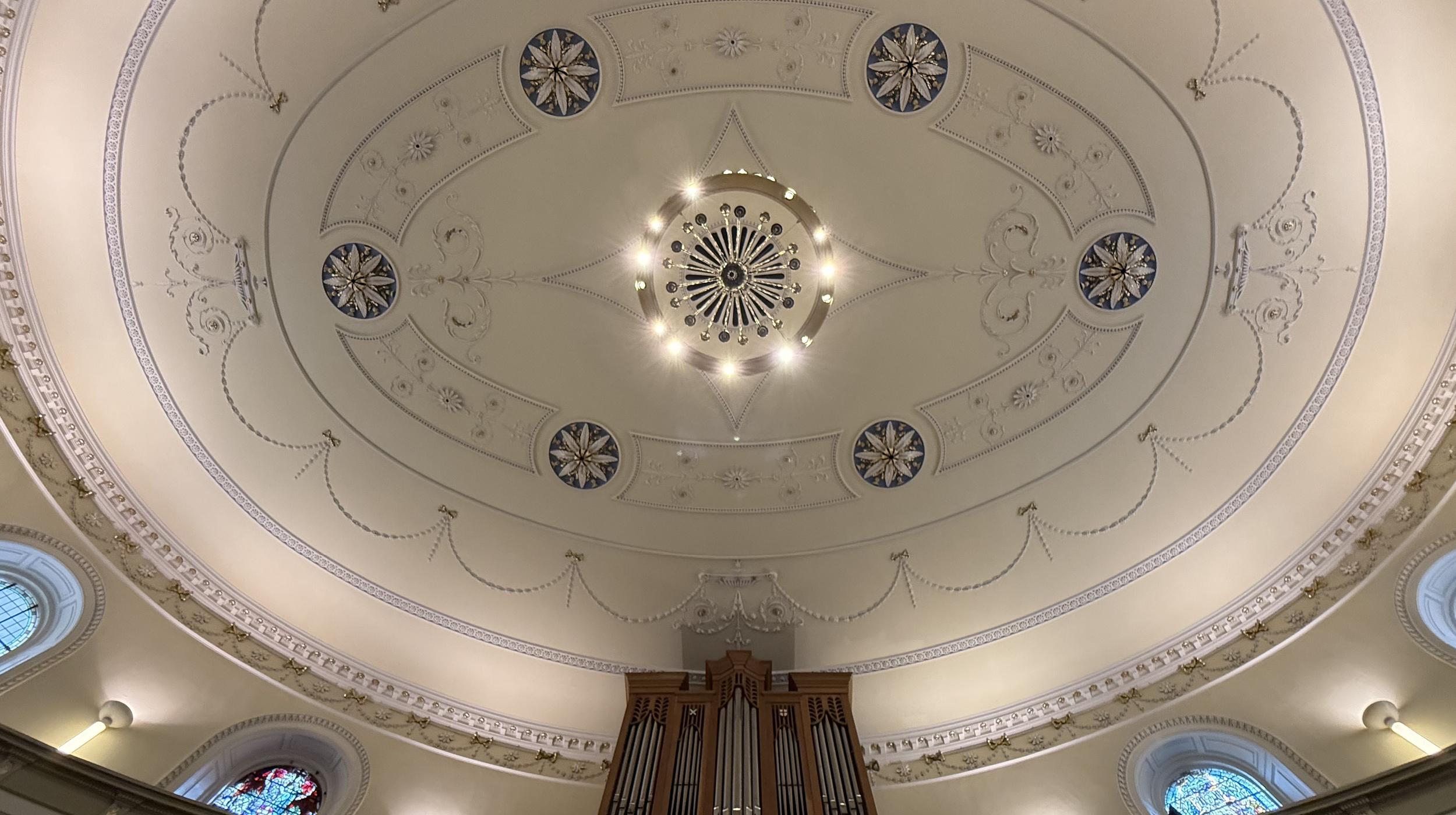 Interior view of a church ceiling featuring ornate moldings, stained glass windows, and a grand chandelier at the center.