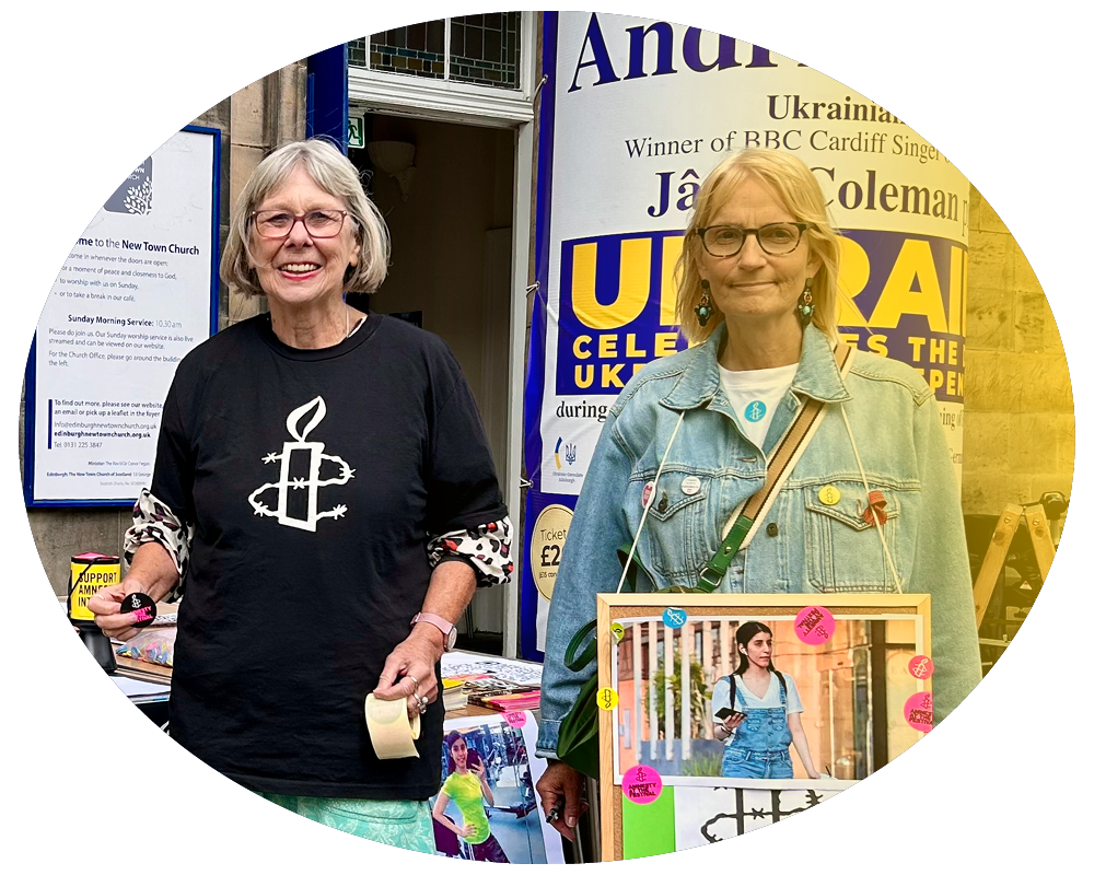 Two women standing outdoors at an event, one wearing a black shirt with a candle and cross symbol, the other in a denim jacket with a framed picture hanging around her neck.