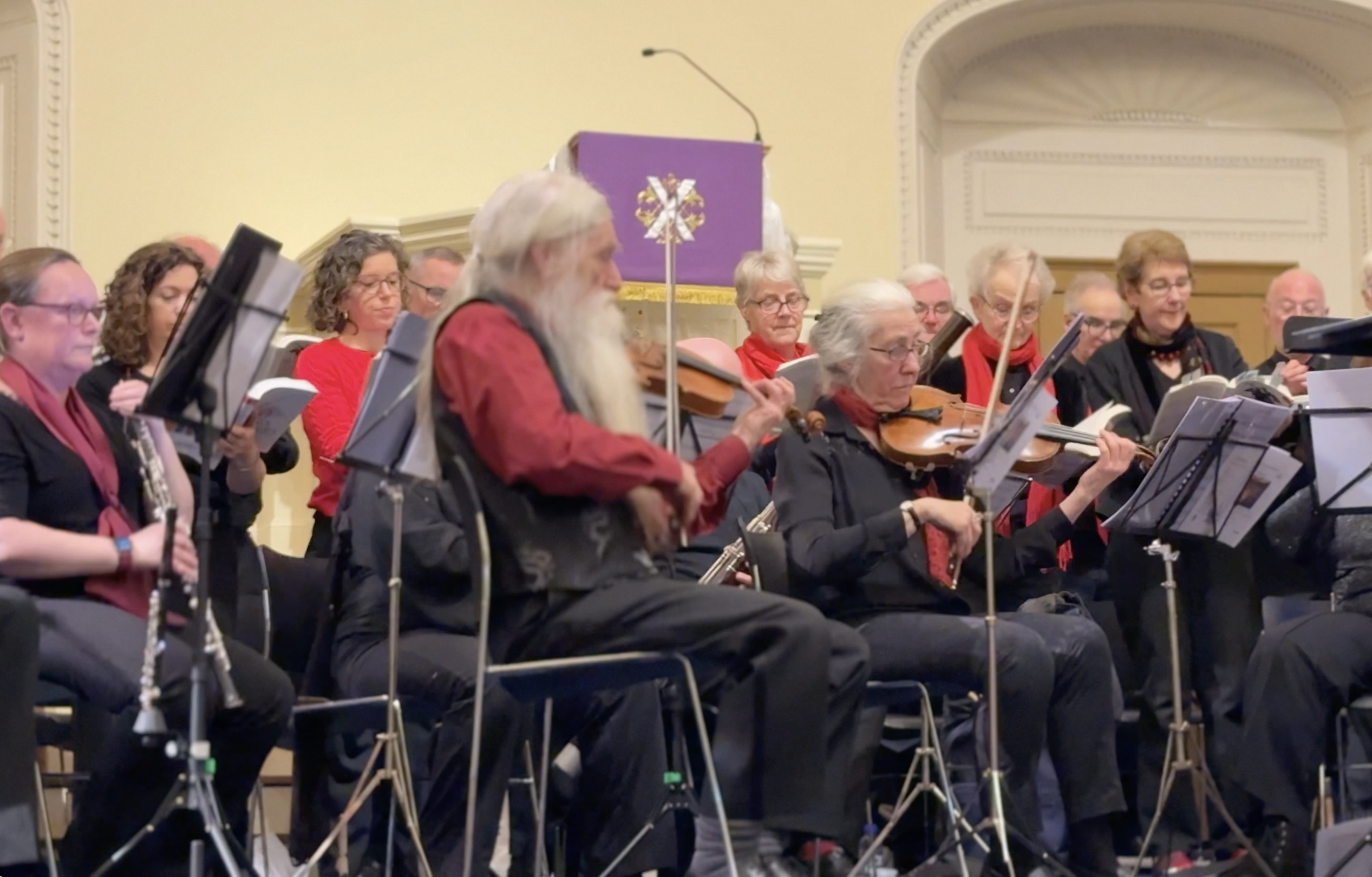 Group of elderly musicians playing in an indoor concert hall with music stands and chairs.