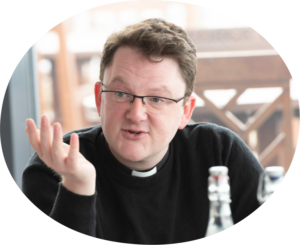 A man with glasses and a clerical collar is sitting at a table, talking, with bottles in front of him.