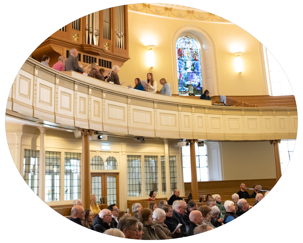A congregation seated inside a church with a pipe organ, stained glass windows, and a balcony with standing people.