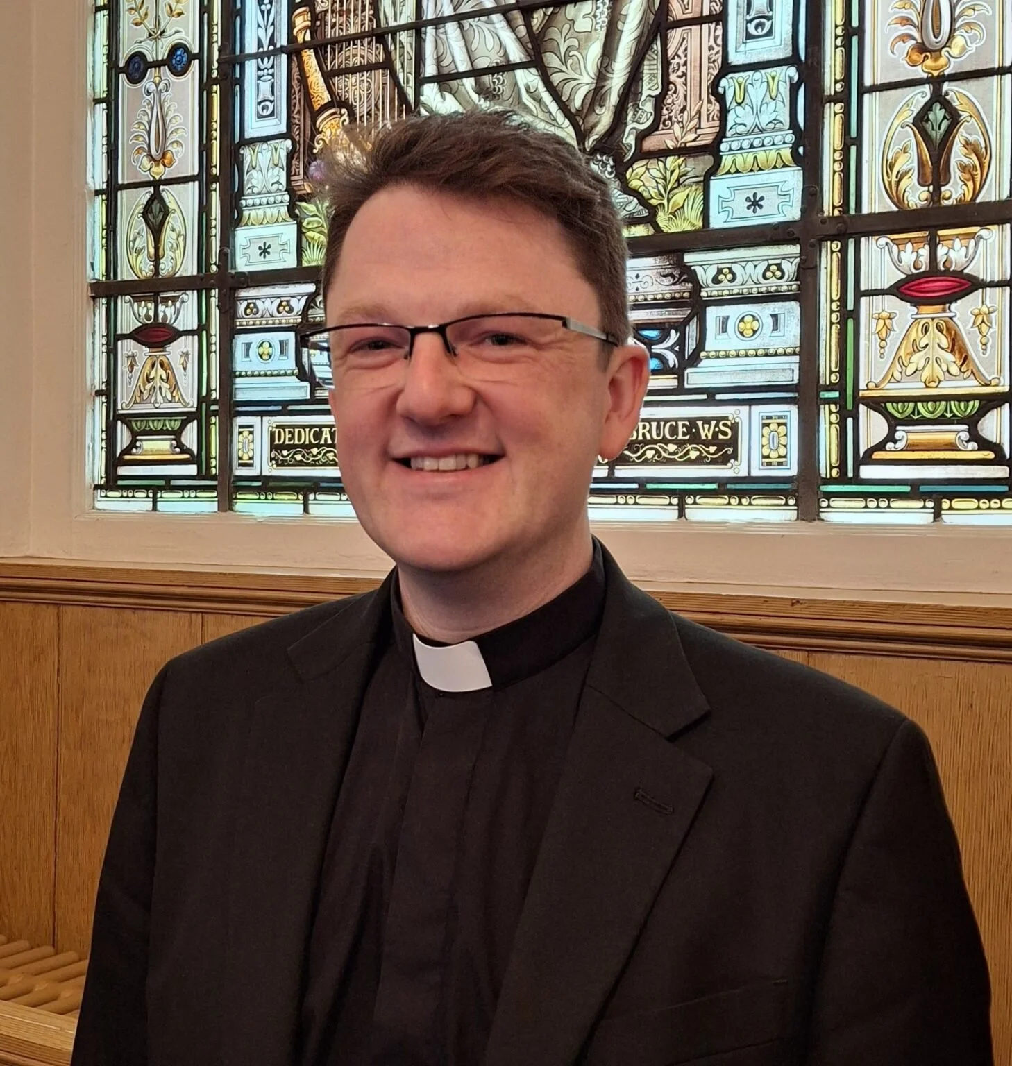 A smiling man dressed as a priest wearing glasses, a black suit, and a clerical collar, standing in front of a decorative stained glass window.