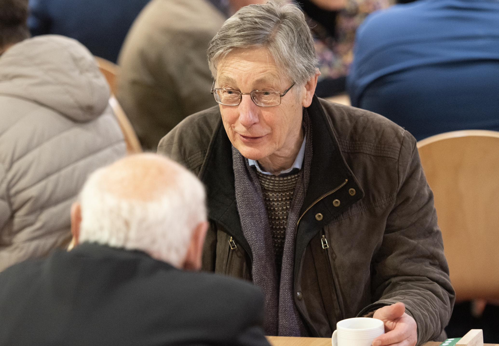 An older man with gray hair and glasses sitting at a table, engaged in conversation with someone, in a crowded indoor setting.