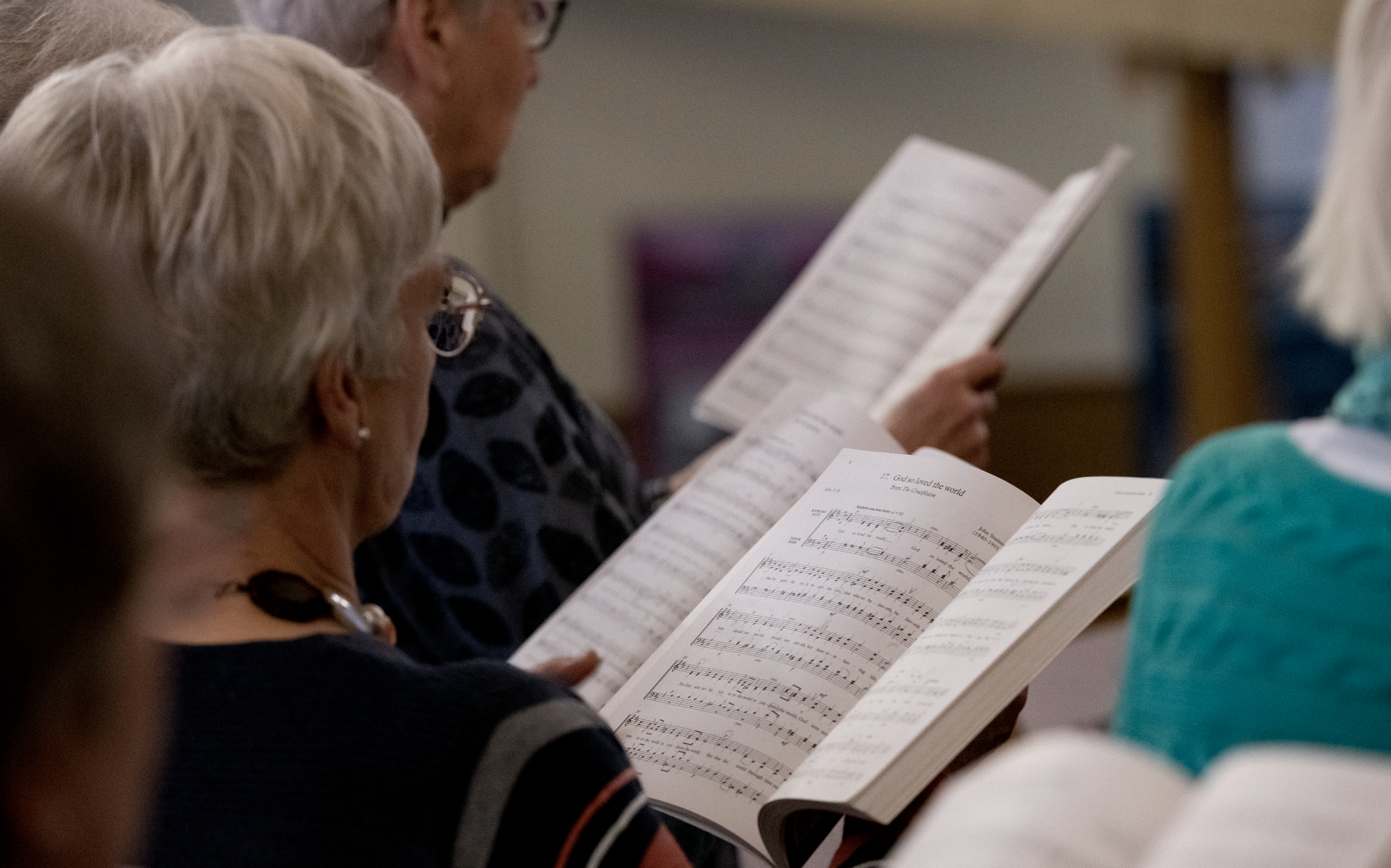 Group of people sitting in a church, singing and holding hymn books, with music sheets visible on the pages.