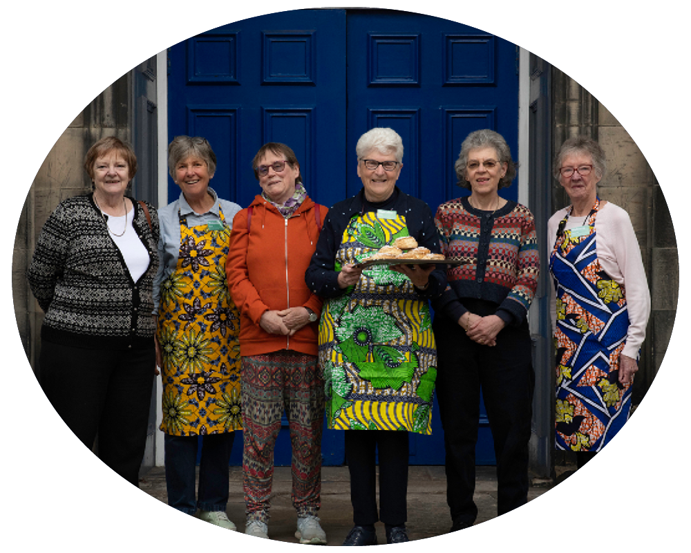 Six elderly women standing in front of a bright blue door, one holding a tray of food, all wearing colorful and patterned clothing.