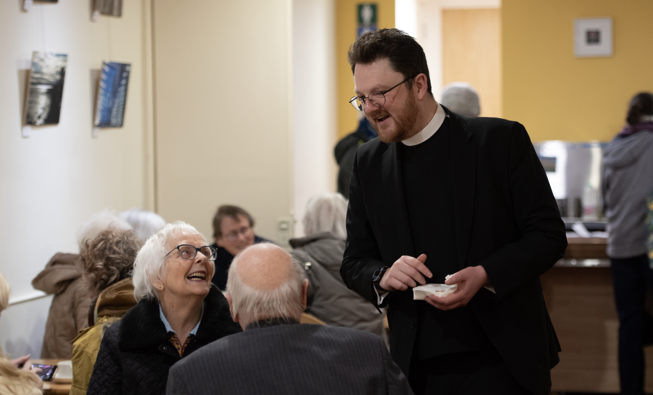 A priest in black clerical attire with glasses smiling and talking to an elderly woman with white hair and glasses in a crowded room, surrounded by other elderly people engaged in conversation.
