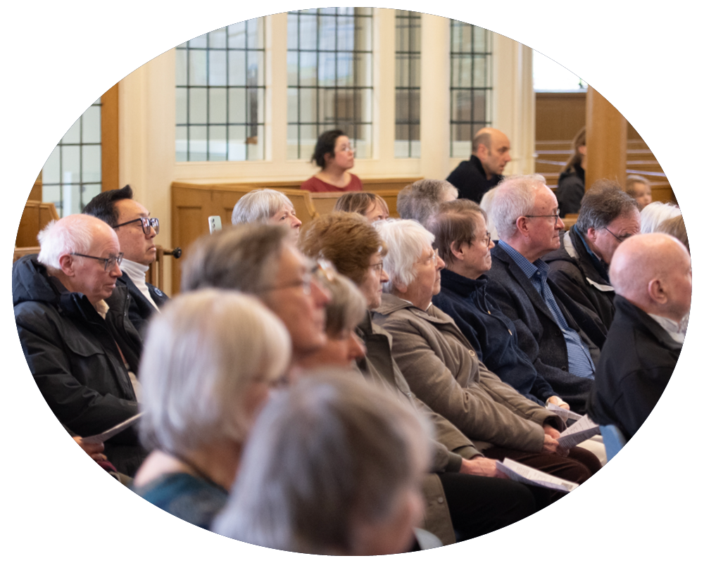 Group of older adults and a few younger individuals seated attentively at an indoor event in a room with large windows.