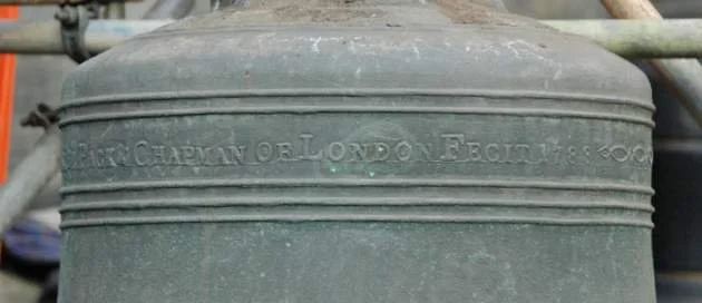 Close-up of an old, green metal bell with inscription reading 'Arthur Chapman of London Fecit 1895'.