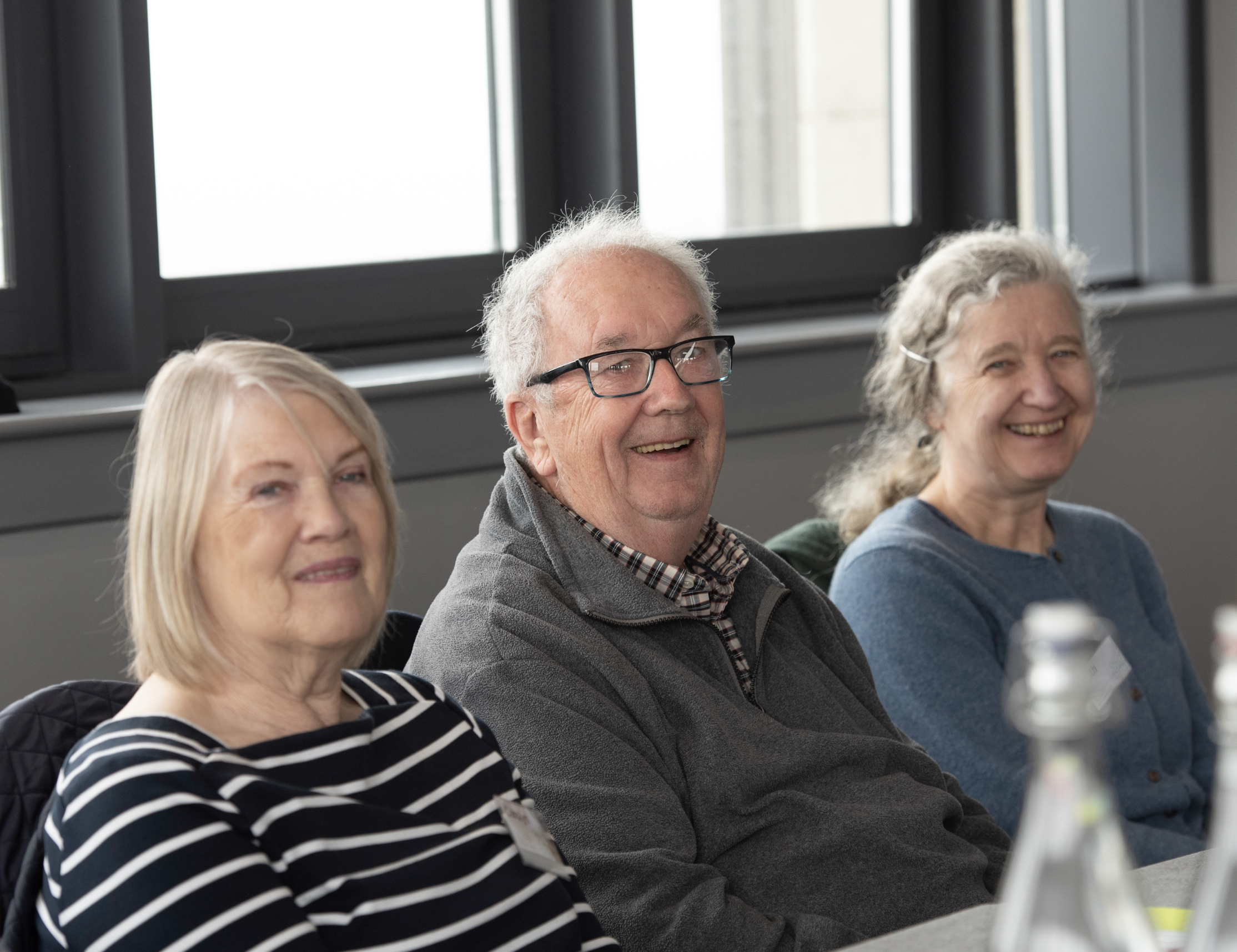 A group of elderly people sitting in a row, smiling during an indoor event, with large windows in the background.