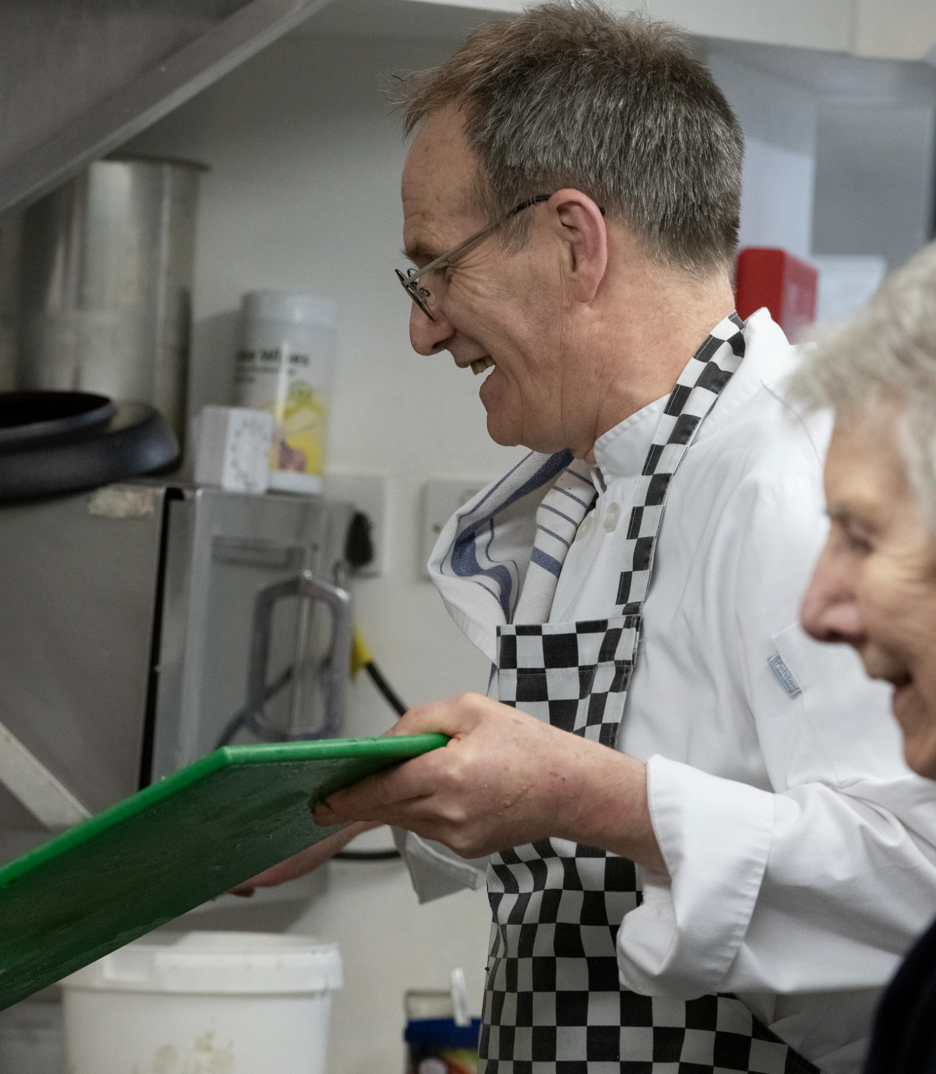 A man wearing glasses and a checkered apron is smiling while looking down at a green tray in a kitchen environment.