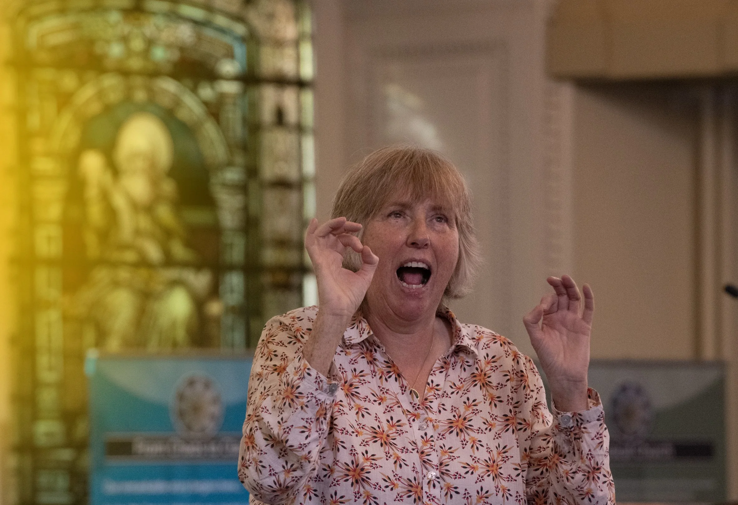 A woman with short blonde hair and freckles, wearing a floral shirt, appears to be speaking or singing, with her mouth open and hands raised near her face. She is in a space with a colorful, blurred background that includes what seems to be a stained glass window.