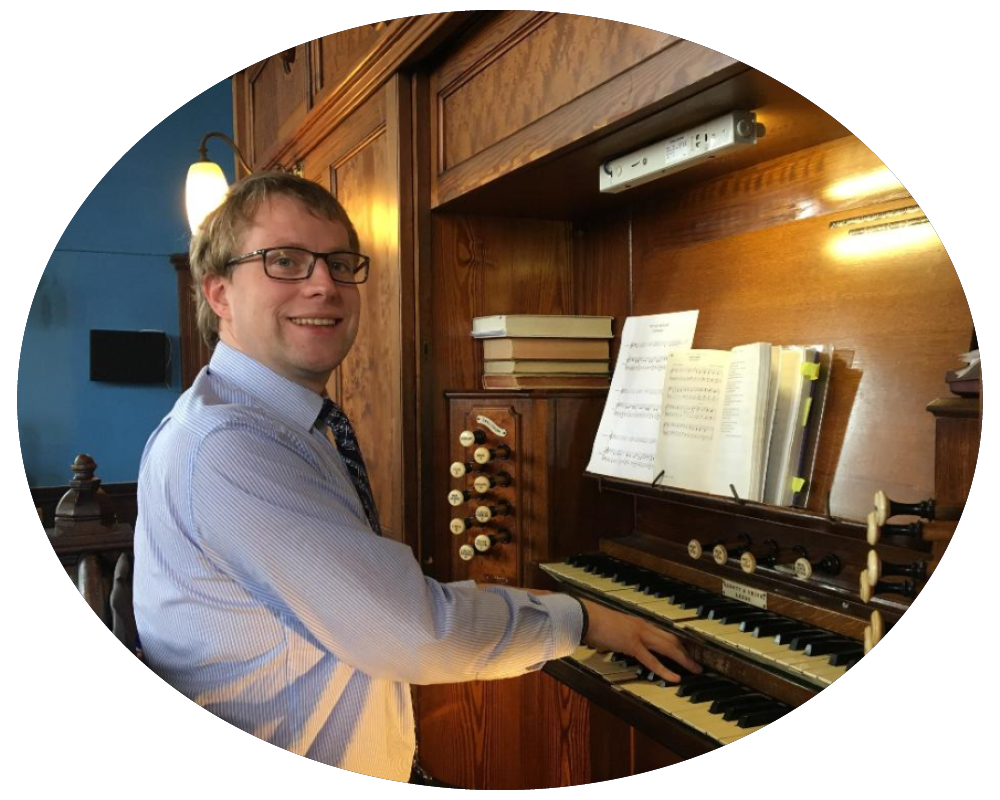 A man in glasses and a dress shirt playing an antique pipe organ with sheet music and books on its wooden console, smiling at the camera.