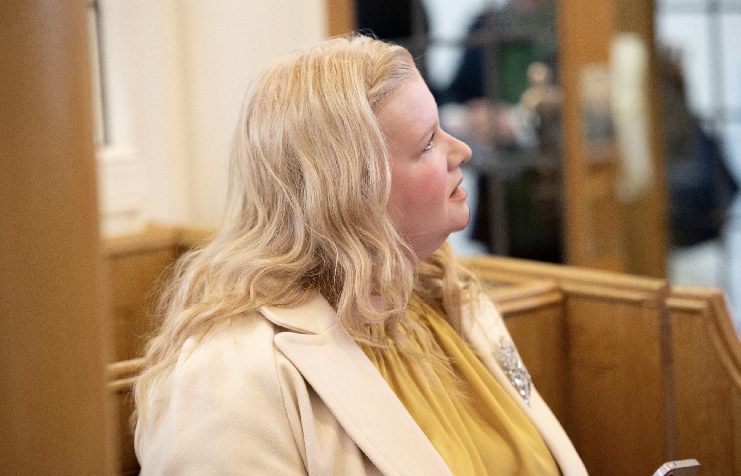 A woman with long blonde hair and a light-colored coat sitting in a wooden pew in a church or courtroom, looking attentively to her right.