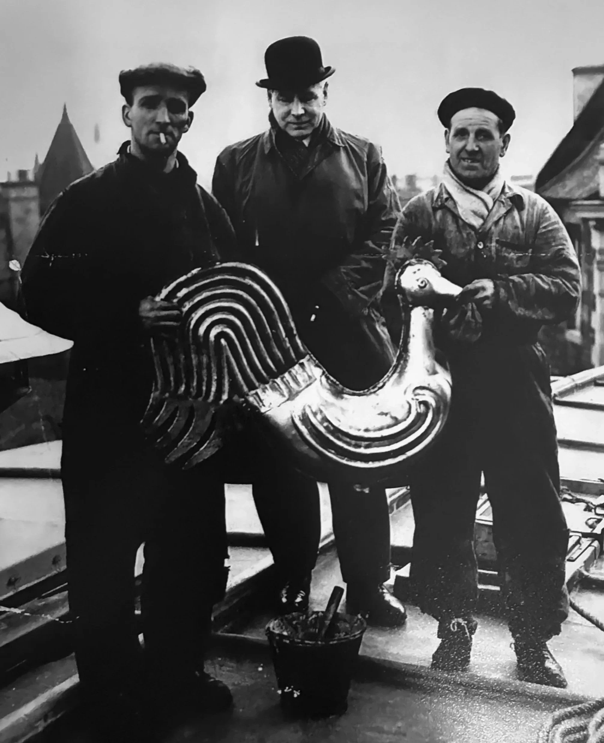 Black and white photo of three men standing outdoors, dressed in vintage clothing. The man on the left has a cigarette in his mouth, wears a dark shirt, and striped pants. The man in the middle wears a hat and a leather jacket. The man on the right wears a beret and a scarf, and holds a toy horse.