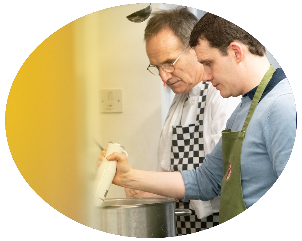 A man and a woman cook together in a kitchen. The man is stirring a pot while the woman holds a handheld blender.