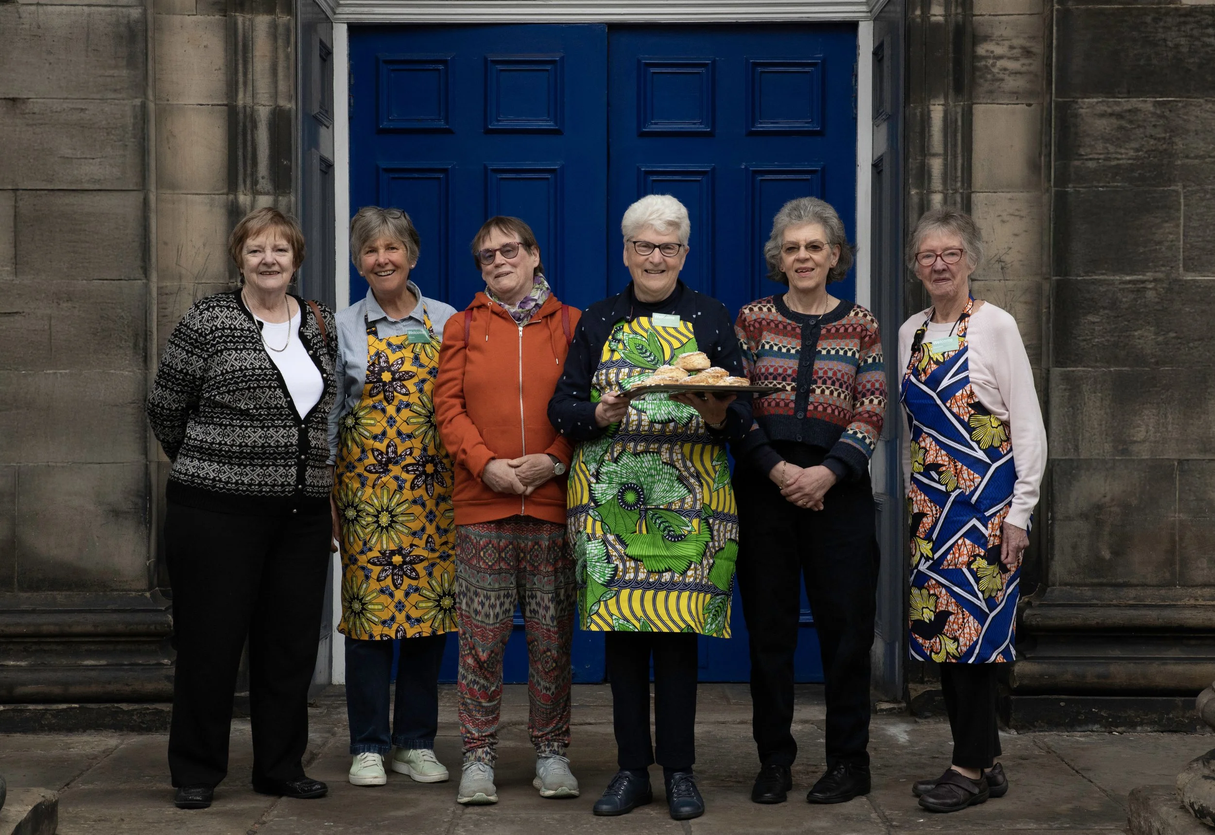Six older women standing in front of a stone building with a large blue door, two of the women are wearing colorful floral aprons, and one woman in the middle is holding a tray of baked goods.