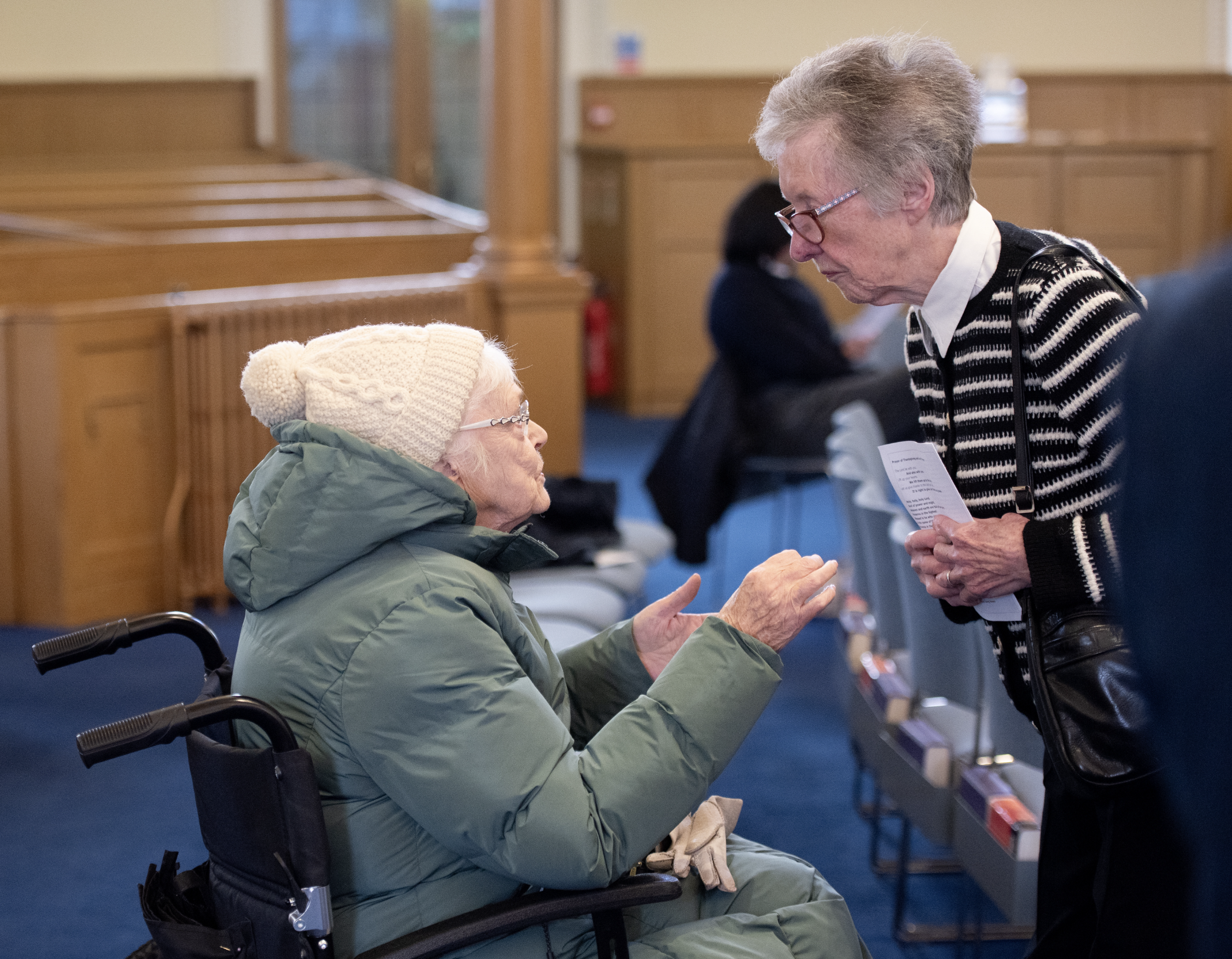An elderly woman with glasses and a white knit hat, sitting in a wheelchair, talking with a woman with gray hair and glasses, who is leaning over while holding a paper.
