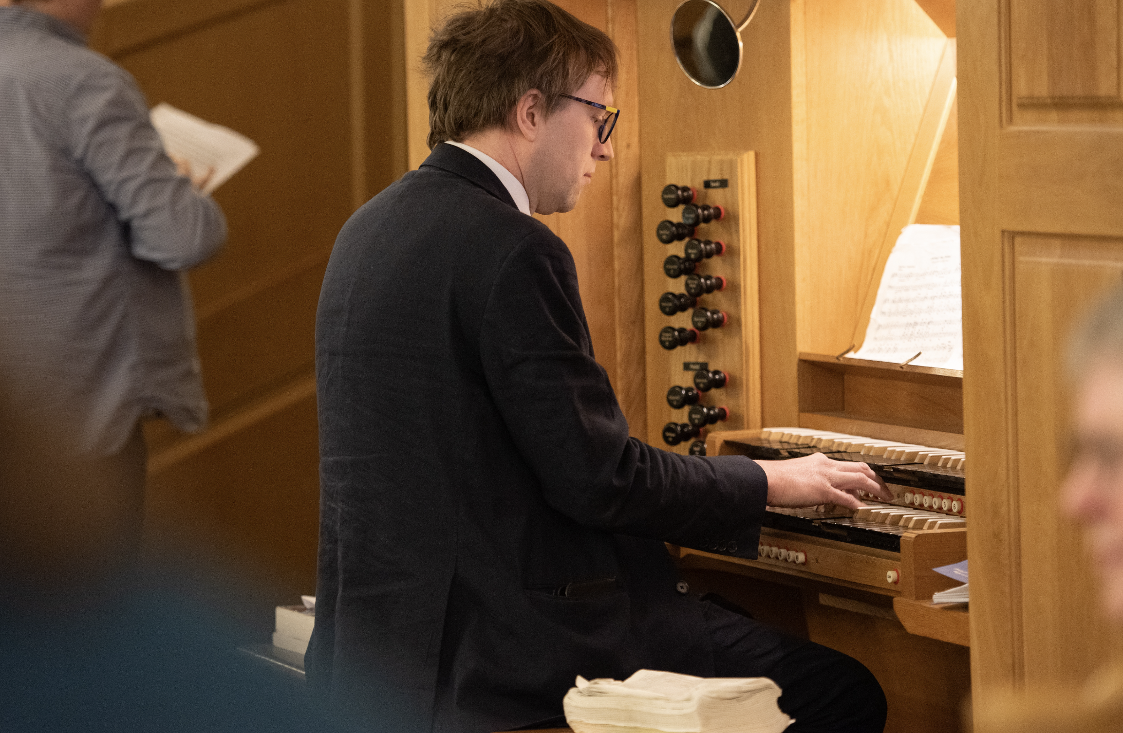 A man wearing glasses and a dark jacket playing an organ in a wooden-paneled room, with sheet music in front of him.