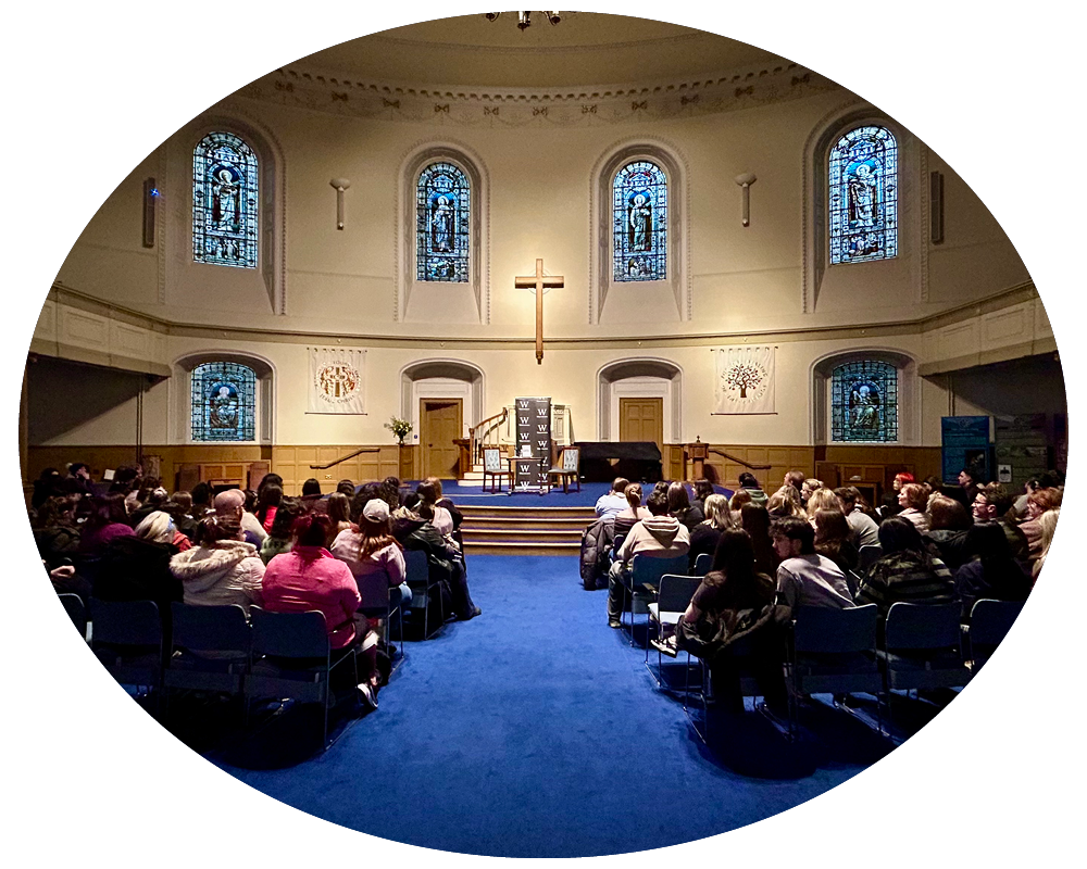 Inside a church with a congregation seated facing the altar, which features a large cross. The church has stained glass windows with religious images and a blue carpeted floor.