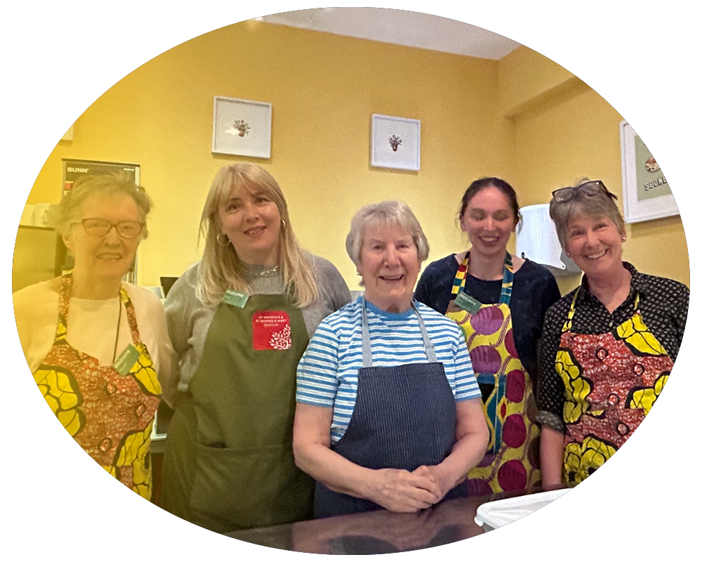 Six women standing together in a room, wearing aprons with colorful patterns, smiling at the camera.