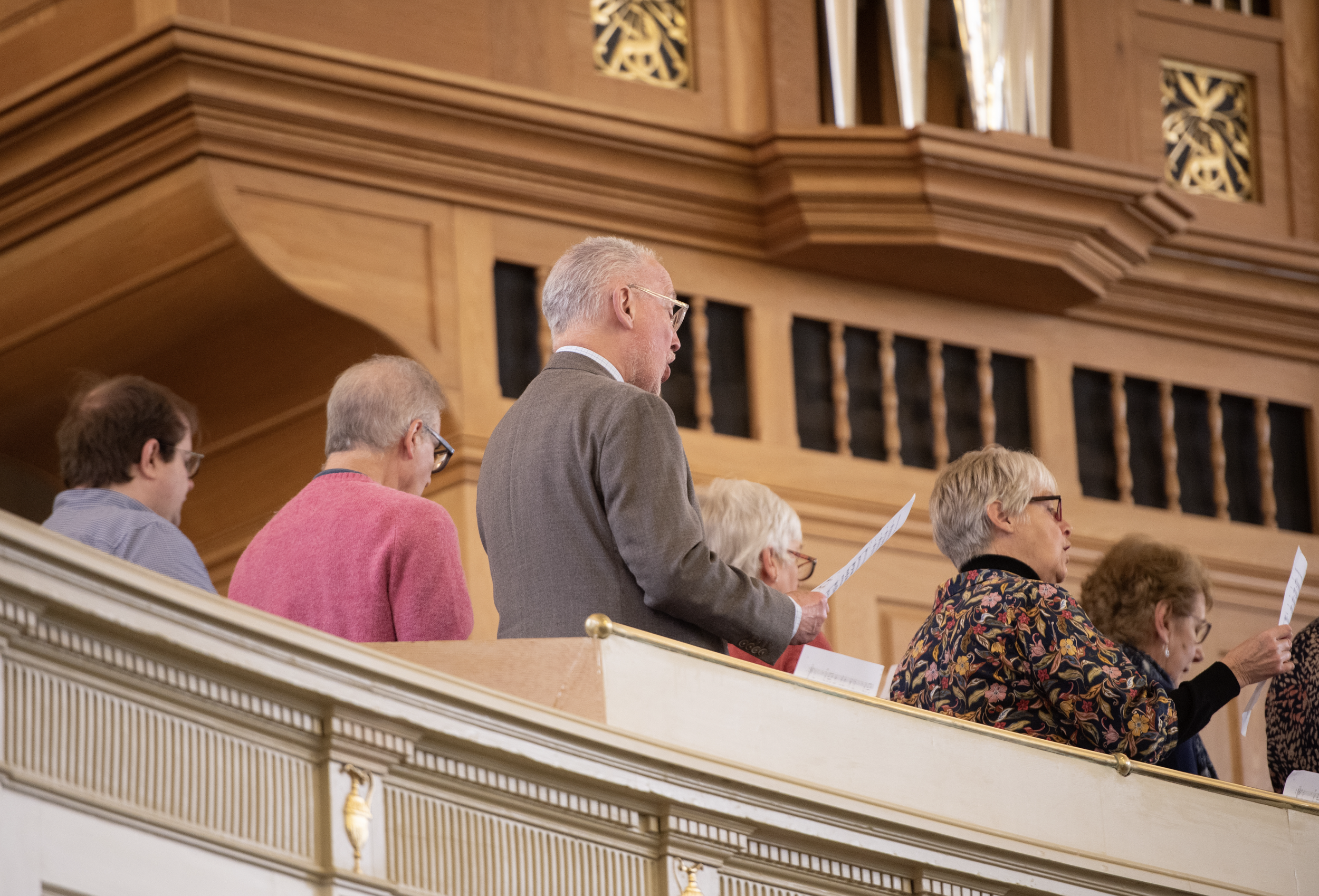 People standing on a balcony with wooden paneling, participating in a formal event, some holding papers, in a building with ornate decorative elements.