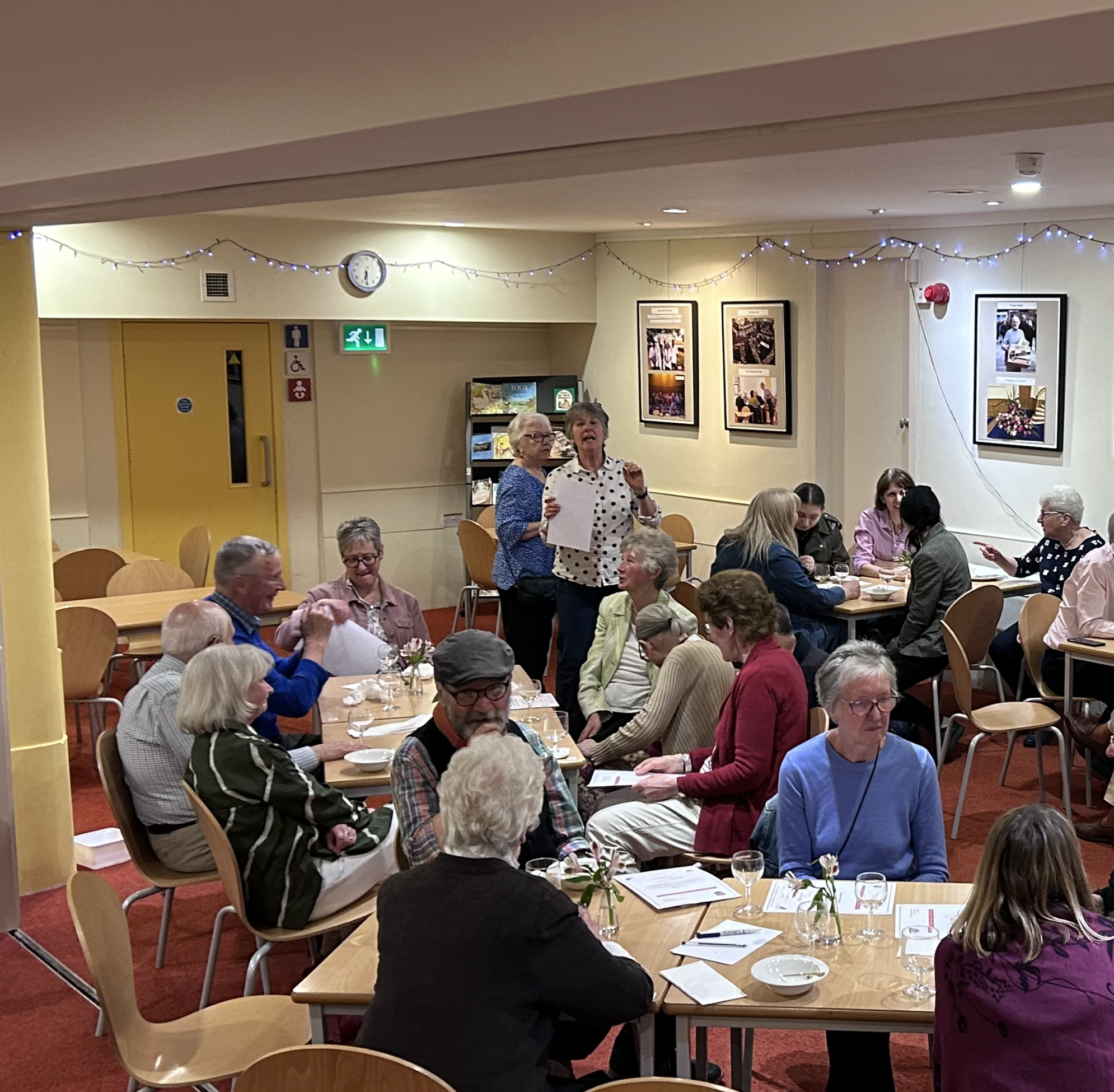 A gathering of older adults in a restaurant or community center, with people sitting at tables, eating, drinking, talking, and some standing and chatting. The setting has decorated walls with framed pictures and string lights, and a clock on the wall showing early evening.