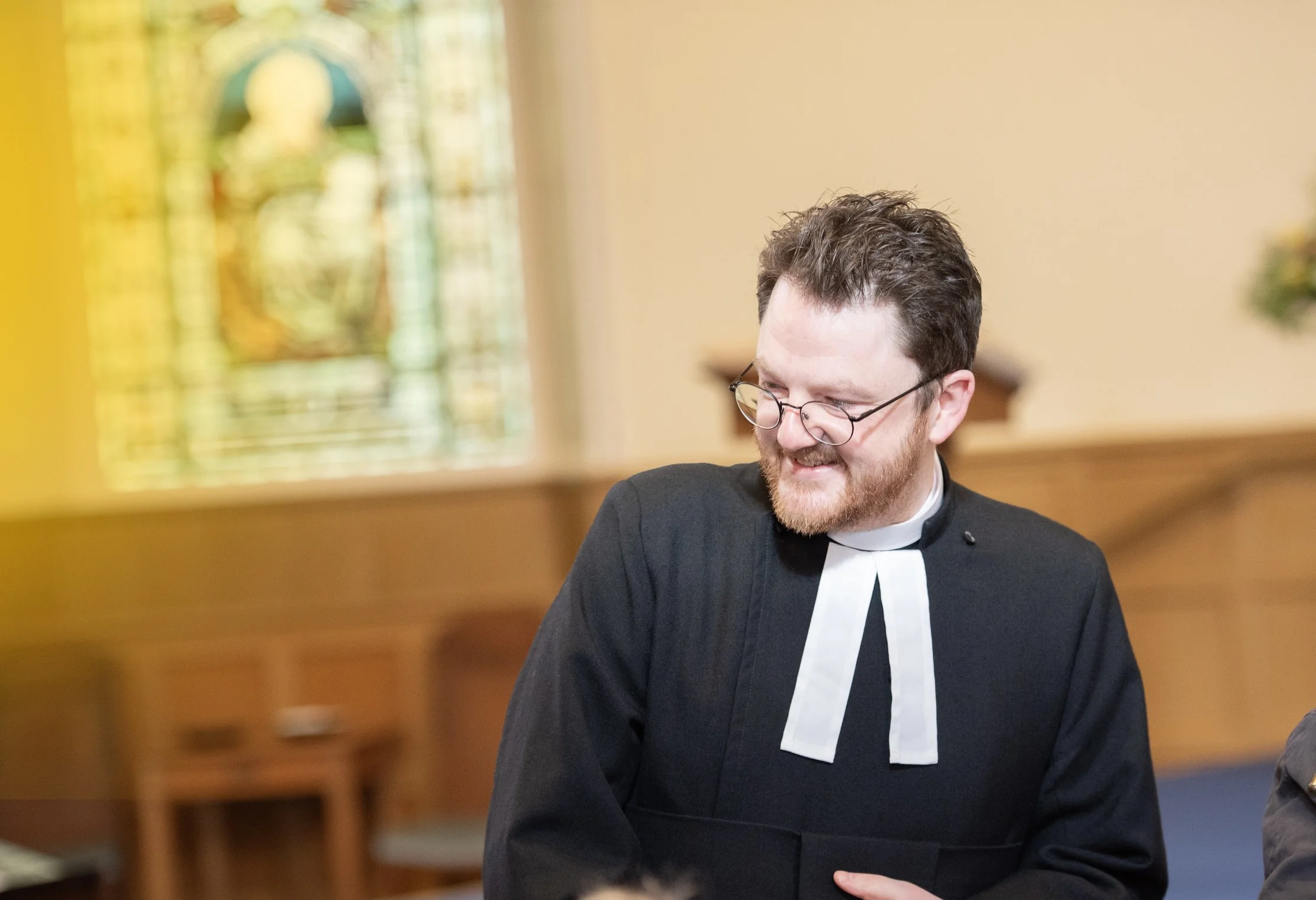 A man dressed as a priest, with glasses and a beard, smiling and looking to his left, inside a church or chapel with a stained glass window in the background.
