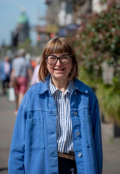 Smiling woman with glasses wearing a blue jacket and striped shirt, standing outdoors on a busy street with people and buildings in the background.