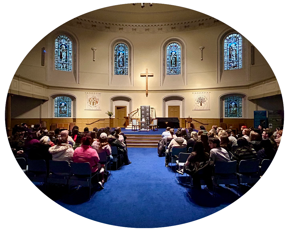 A church interior with a congregation seated facing the altar, which has a cross mounted on the wall above a small stage with chairs and a piano. The church has stained glass windows and religious symbols on the walls.