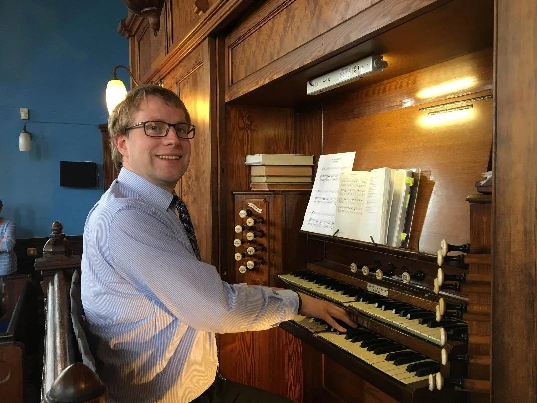 A man with glasses and a striped shirt playing an organ or a large keyboard in a church or chapel.
