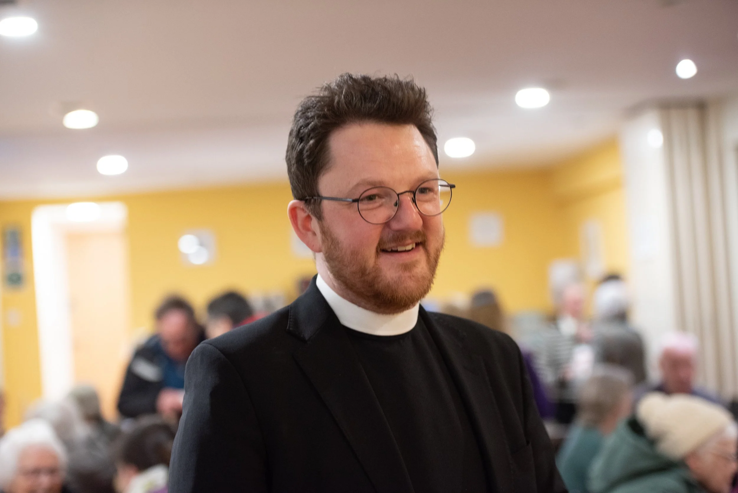 A man with glasses and a beard is smiling, wearing a black suit and clerical collar, in a room with yellow walls and ceiling lights, with people seated and standing in the background.