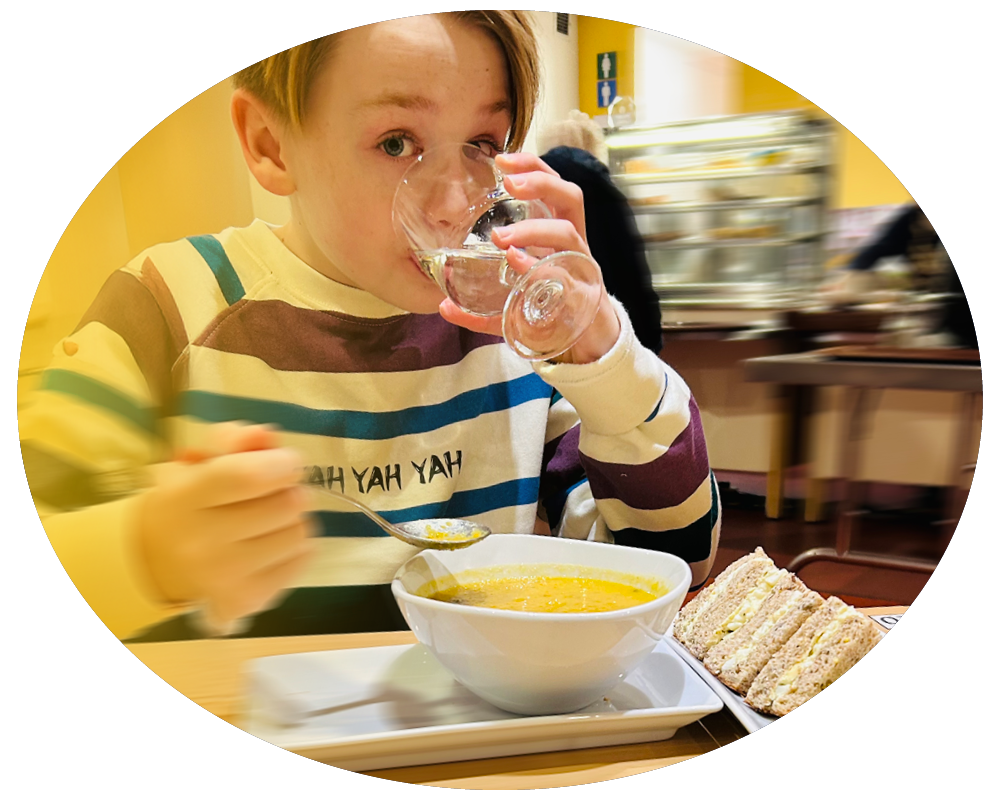 A young boy drinking water from a glass at a table with a bowl of soup and a sandwich.