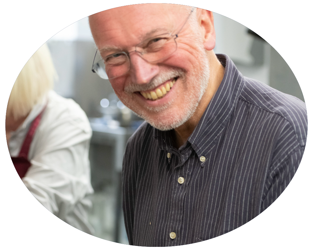 A smiling older man with glasses and a beard, wearing a dark striped button-up shirt, in a kitchen setting.