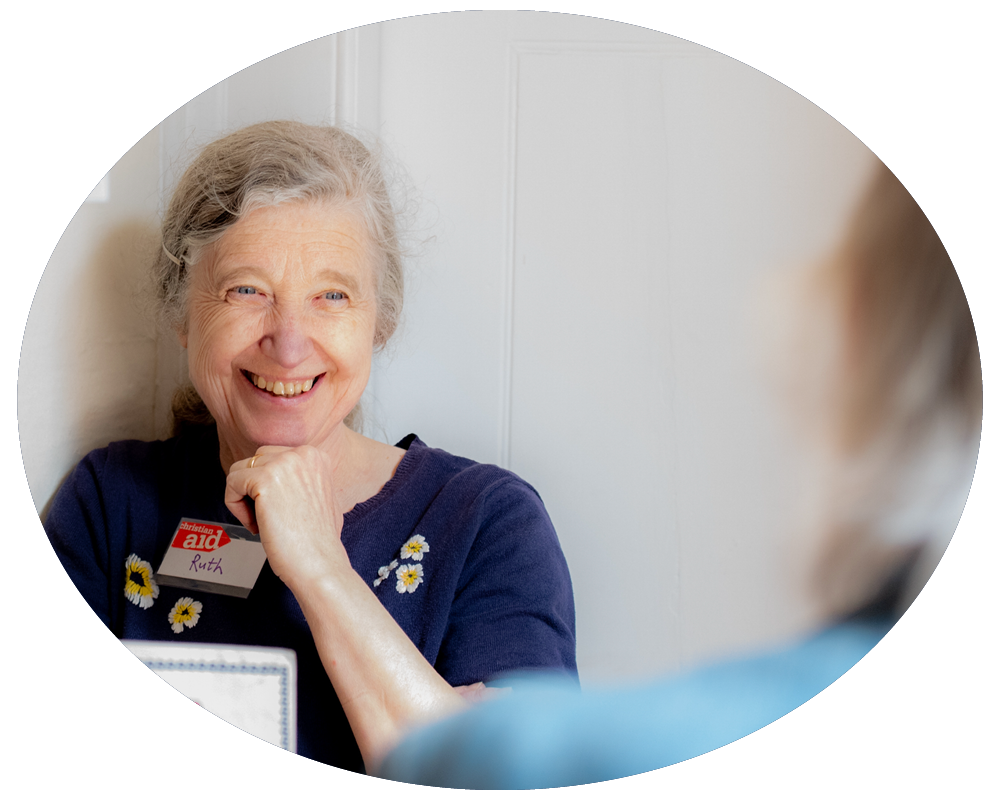 Elderly woman with gray hair smiling, wearing a dark blue shirt with flower embroidery, and a badge that reads 'Ruth', talking to a person who is partially visible in the foreground.