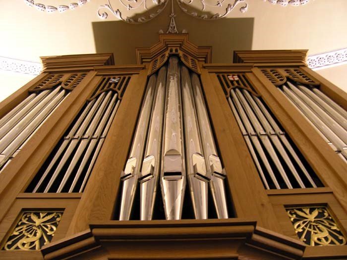 A close-up of a wooden pipe organ with metal pipes, viewed from below, with decorative elements and a ceiling in the background.