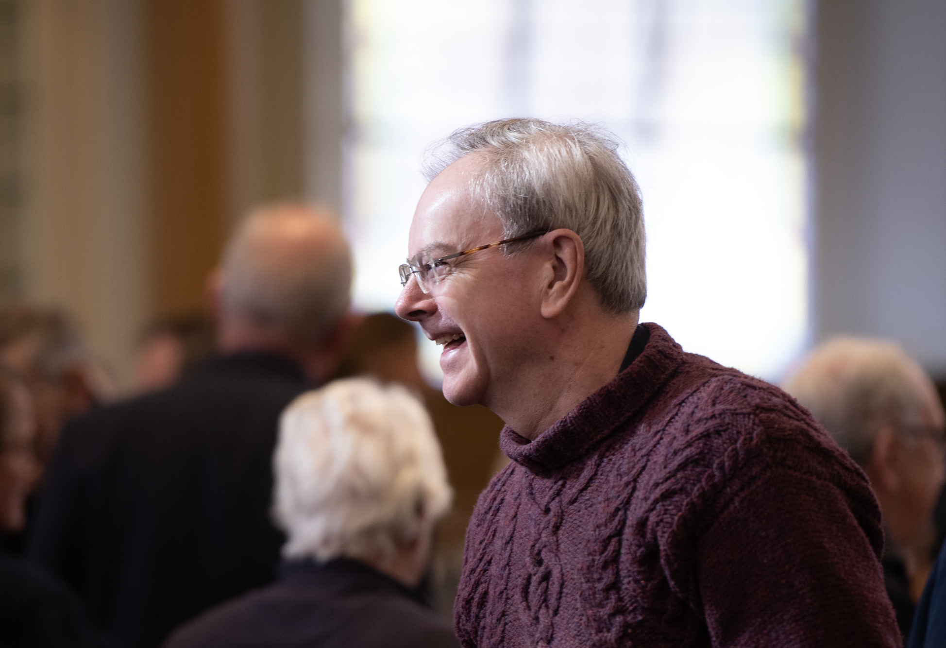 A smiling man with glasses and light-colored hair wearing a maroon sweater, engaged in conversation at a gathering with blurred people in the background.