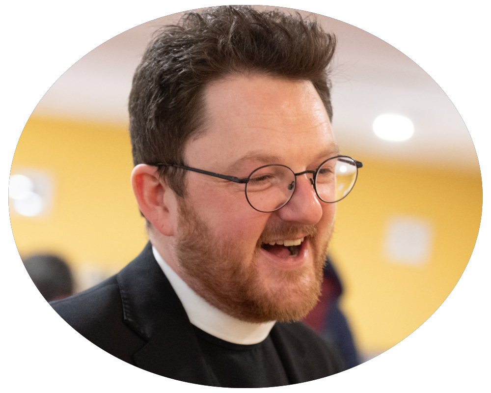 A man with glasses and a beard, possibly a priest, smiling and wearing a clerical collar in an indoor setting.