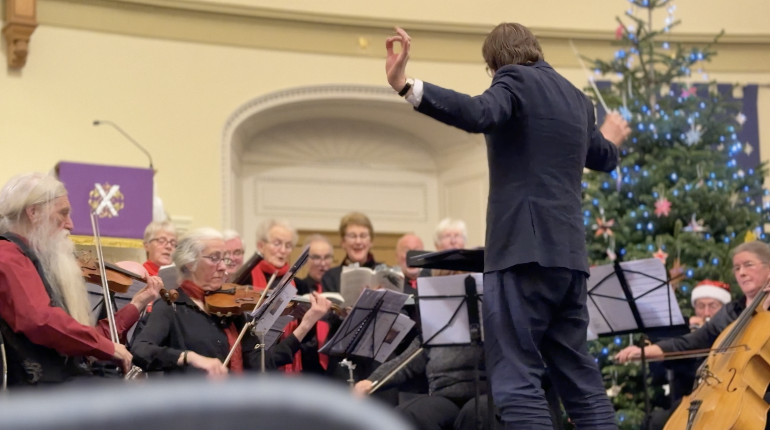 Conductor leading a choir and orchestra performing holiday music near a decorated Christmas tree.