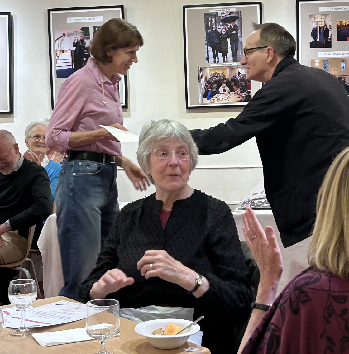 A woman wearing a black top and glasses is talking, while another woman with short gray hair is sitting at a table with a bowl of food and glasses of water. A man in a dark jacket is leaning over, shaking hands or greeting the woman with short hair. Several people are in the background, some smiling or clapping, in a dining or event hall decorated with framed photos on the wall.